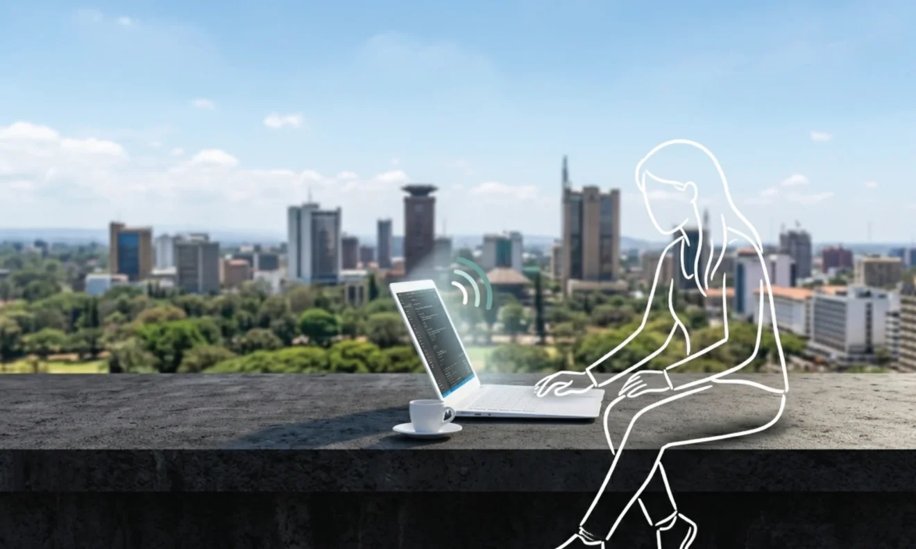 A slim open laptop on a dark concrete rooftop surface in Nairobi, the full city skyline stretching luminously behind it under a bright midday sky